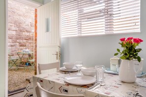 Un coin repas avec une table dressée pour deux personnes, un pichet blanc orné de fleurs et une fenêtre avec des stores.