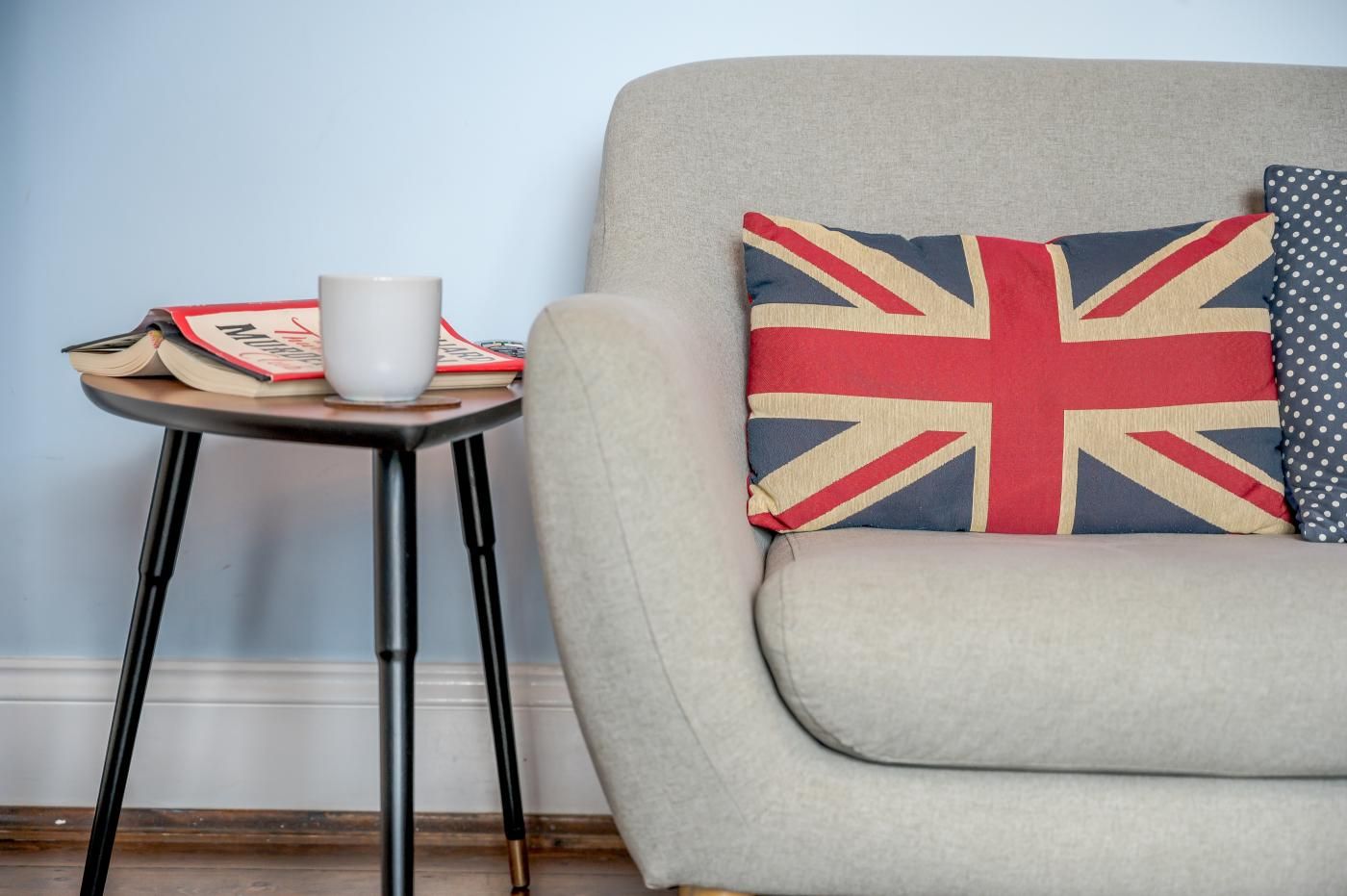 A room with a grey armchair, a small table with a white mug, books, and a pillow with a Union Jack design.