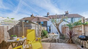 An outdoor patio with a wooden deck, yellow chairs, a table with a drink, and a barbecue grill. - 2 Bed in Whitstable (oc-o32772) (Whitstable)