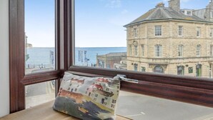 A window seat with a cushion and books, overlooking a coastal town with historic buildings. - 2 Bed in Swanage (oc-wy547) (Swanage)