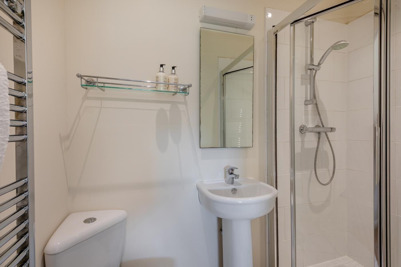 A bathroom with a toilet, a sink, a glass shelf with toiletries, a mirror, and a glass-enclosed shower.