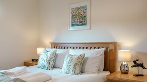 Een net opgemaakt bed met witte lakens, twee nachtkastjes met lampen, een houten hoofdbord en een lijst met bloemkunstwerk erboven.