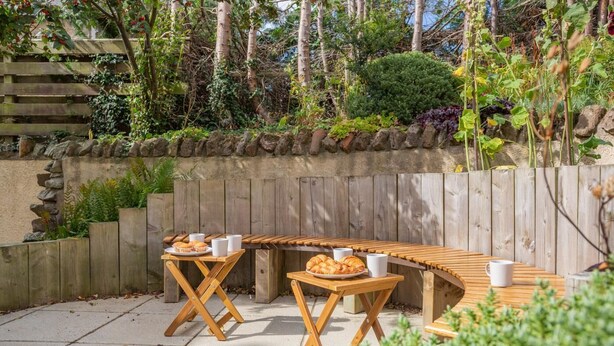 Een buitenplaats met een eethoek bestaande uit een gebogen houten tafel en stoelen, omgeven door een stenen muur en diverse planten.