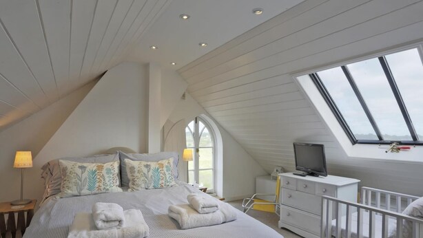 Ein Schlafzimmer mit schrägen Wänden, einem Bett mit gestreifter Bettwäsche, einem Fernseher, einem Schreibtisch und einem Fenster mit Blick.