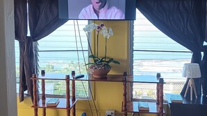 Una habitación con televisión montada en la pared, ventilador de techo, ventana con cortinas, mesa de madera con planta en maceta y una mesita con lámpara.