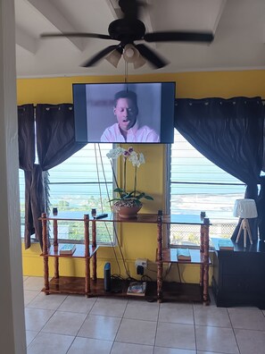 A room with a television mounted on the wall, a ceiling fan, a window with curtains, a wooden table with a potted plant, and a small table with a lamp.