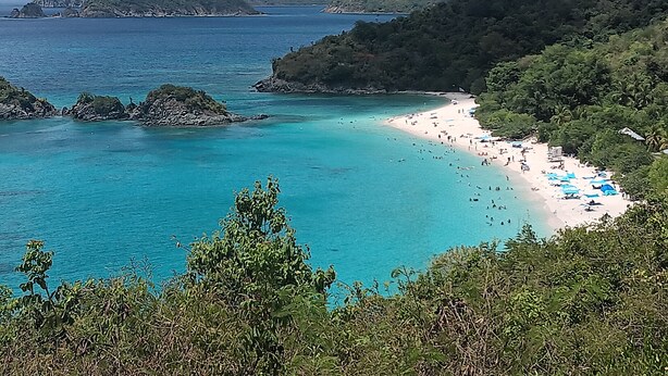 En strand med kristallklart turkosfärgat vatten, en sandstrand och frodiga, gröna kullar i bakgrunden.