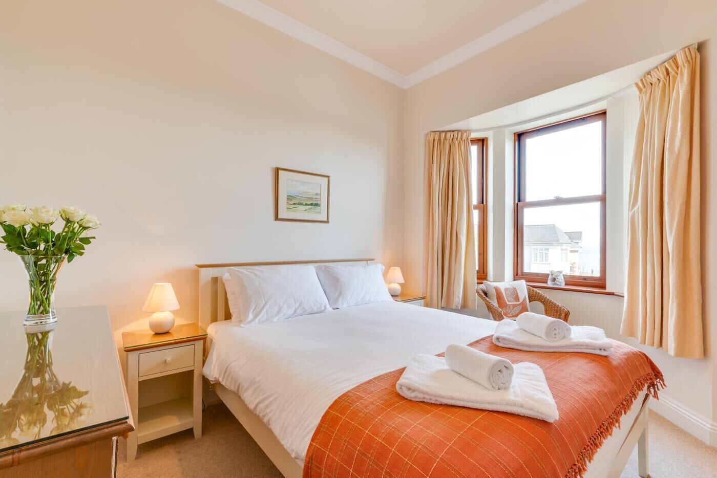 A hotel room with a bed, a nightstand, a lamp, a window with curtains, and a vase with flowers.
