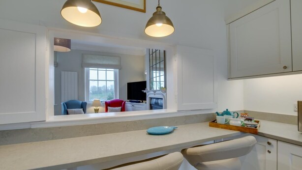 Una cocina con encimera blanca, dos taburetes de bar, un plato azul y una ventana que ofrece vistas a una silla azul y una silla roja.