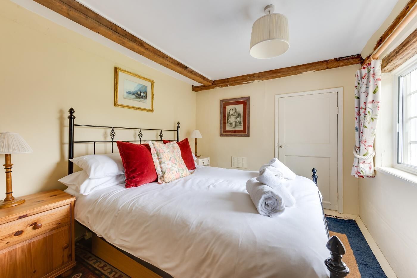 A neatly made bed with white linens and red pillows, a wooden nightstand with a lamp, and a window with floral curtains.