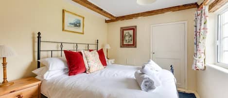 A neatly made bed with white linens and red pillows, a wooden nightstand with a lamp, and a window with floral curtains.