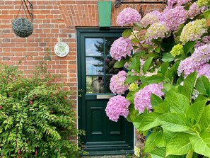 A brick building with a green door, a hanging sign, and pink hydrangeas in front. - 3 Bed in Saxmundham (oc-tnk) (Dunwich)