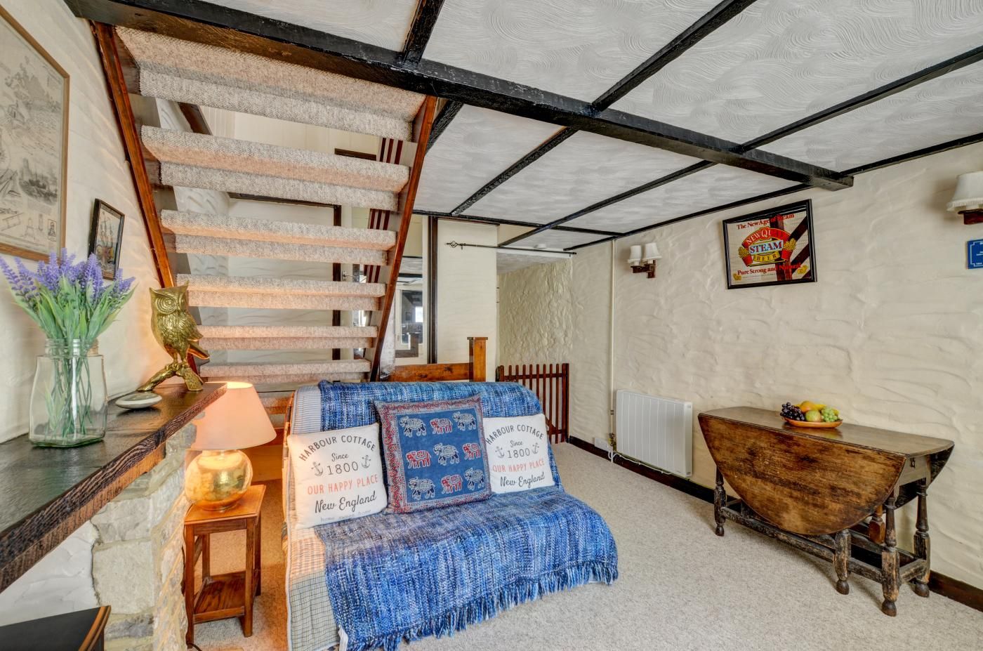 Une pièce chaleureuse avec un escalier en bois, un banc bleu en osier, une table en bois sur laquelle sont disposés des fruits, et une lampe fixée au mur.