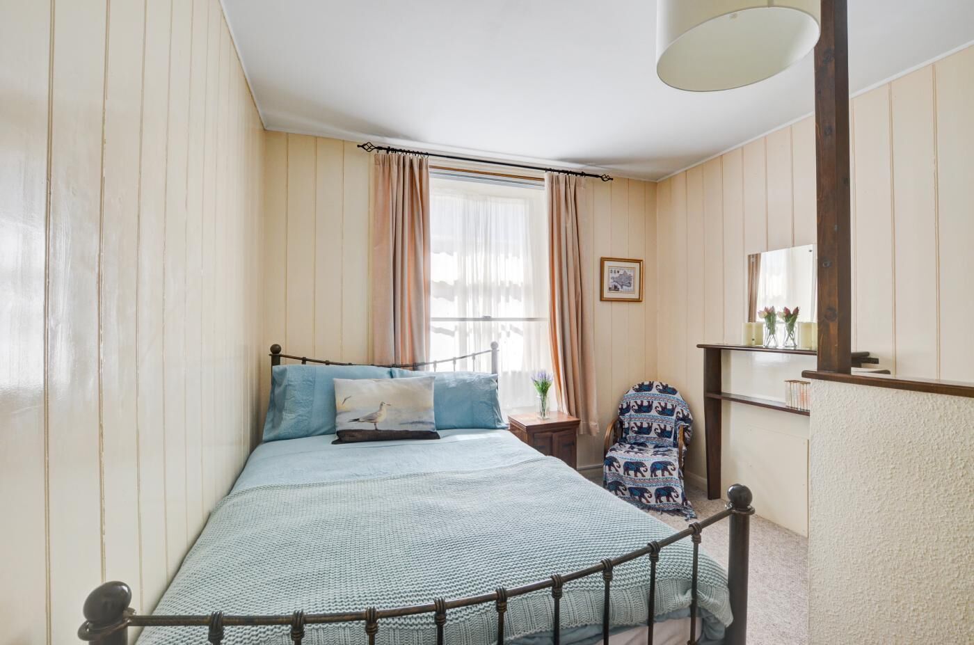 Een slaapkamer met een bed, een stoel, een nachtkastje, een spiegel en een raam met gordijnen.