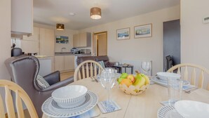 Un coin repas avec une table dressée pour quatre personnes, des chaises, un panier de fruits et une cuisine en arrière-plan.