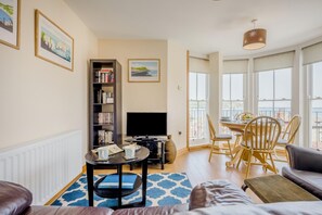 A living room with a sofa, a coffee table, a bookshelf, a flat-screen TV, and a dining area with chairs and a table. - 2 Bed in Swanage (oc-wy513) (Swanage)