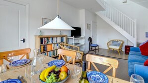 Un comedor con una mesa lista para dos personas, rodeada de sillas de mimbre y un sofá azul. Se ven una estantería y un televisor al fondo.