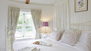 Un dormitorio con cama, almohadas, una mesita de noche con lámpara, una ventana con cortinas, y un cuadro enmarcado en la pared.