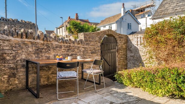 A stone wall, a wooden table with chairs, and a stone archway. - 3 Bed in Swanage (oc-wy272) (Swanage)