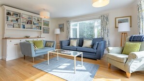 Ein helles Wohnzimmer mit einem blauen Sofa, einem grauen Sessel, einem Glastisch und einem Bücherregal voller Bücher und Dekoration.