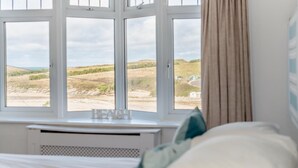 A bedroom with a large window offering a view of a beach and distant landscape. - 5 Bed in Newquay (oc-c33035) (Newquay)
