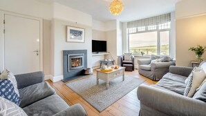 A modern living room with a fireplace, grey sofas, a coffee table, and a large window with a view. - 5 Bed in Newquay (oc-c33035) (Newquay)