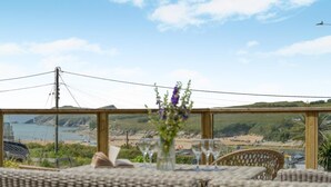 A dining area with wicker furniture, a table with a vase of flowers, and a scenic view of a beach and cliffs. - 5 Bed in Newquay (oc-c33035) (Newquay)