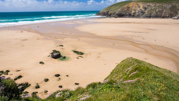 A wide sandy beach with gentle waves, a grassy cliff, and a clear blue sky. - 5 Bed in Newquay (oc-c33035) (Newquay)