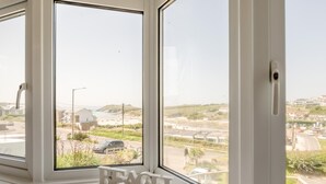 A room with large windows overlooking a beach, a small 'BEACH' sign, and a view of a residential area. - 5 Bed in Newquay (oc-c33035) (Newquay)