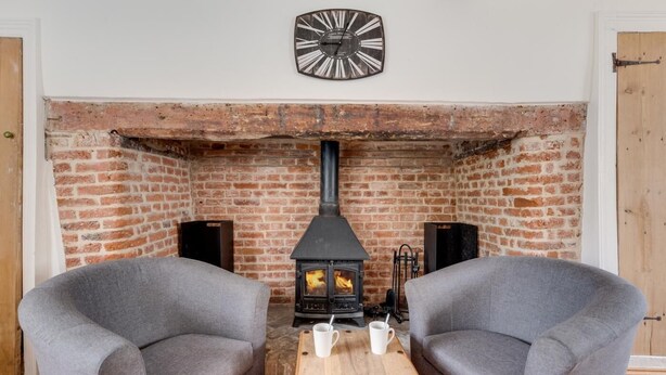 A cozy room with a brick fireplace, two grey armchairs, a wooden table with cups, and a clock on the wall. - 4 Bed in Saxmundham (oc-oth) (Dunwich)