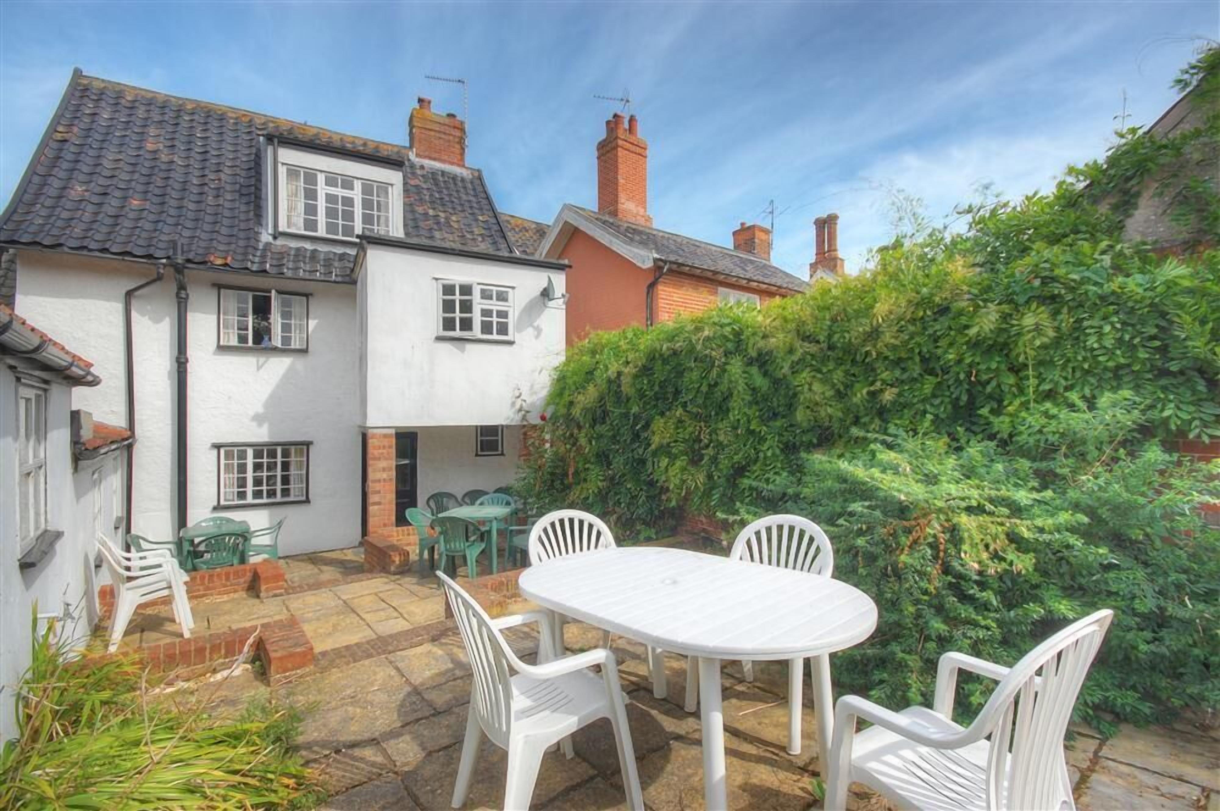 A white house with a tiled roof, a brick chimney, and a garden with a white table and chairs.