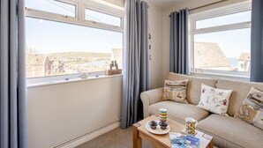 Ein gemütliches Zimmer mit einer Couch, einem Tisch mit Tasse und Gebäck, und Blick auf eine Küstenstadt durch große Fenster.