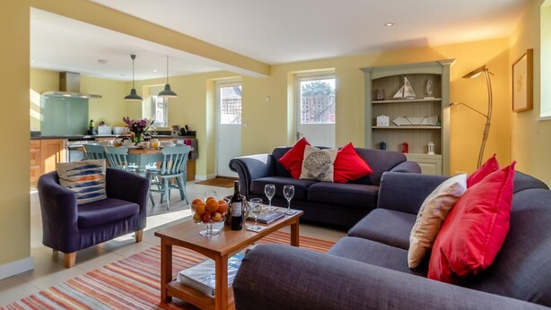 A modern living room with a sofa, armchairs, and a coffee table. There is a kitchen in the background with a dining area. - 3 Bed in Knodishall (oc-wbco) (Knodishall)