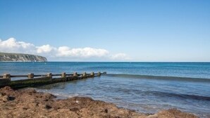 Eine felsige Küstenlinie mit einem hölzernen Steg, der sich in ruhiges, blaues Wasser erstreckt, bewölkter Himmel und eine ferne Klippe.