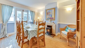 A cozy dining room with a wooden table set for four, a fireplace, and a wicker chair. - 3 Bed in Southwold (oc-sl139) (Southwold)