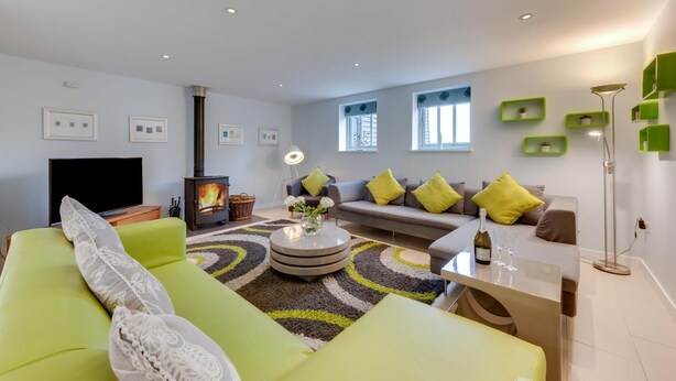 A modern living room with a green sofa, a flat-screen TV, a fireplace, and a dining area with a table and chairs. - 6 Bed in Knodishall (oc-bil) (Knodishall)