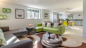 Ein modernes Wohnzimmer mit einem grünen Sofa, einem grauen Sessel und einem Couchtisch mit einem Gesellschaftsspiel. Im Hintergrund ist eine Küche mit einem Essbereich zu sehen.