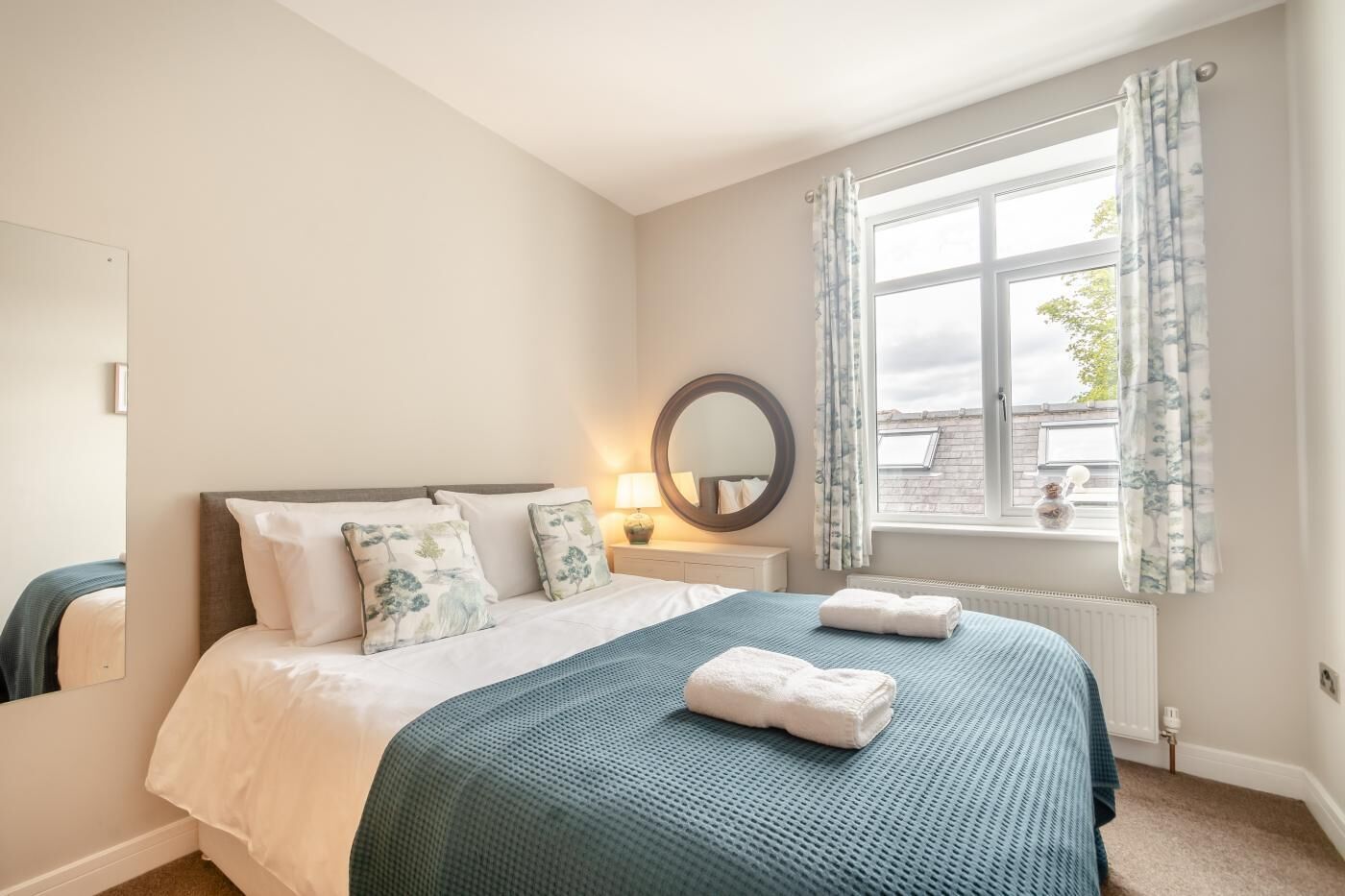 A bedroom with a large bed, a bedside table with a lamp, a window with curtains, and a radiator.