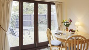 Ein Zimmer mit einer Schiebetür zu einer Terrasse. Dort befindet sich ein Tisch mit Stühlen, eine Vase mit Blumen und eine Lampe.