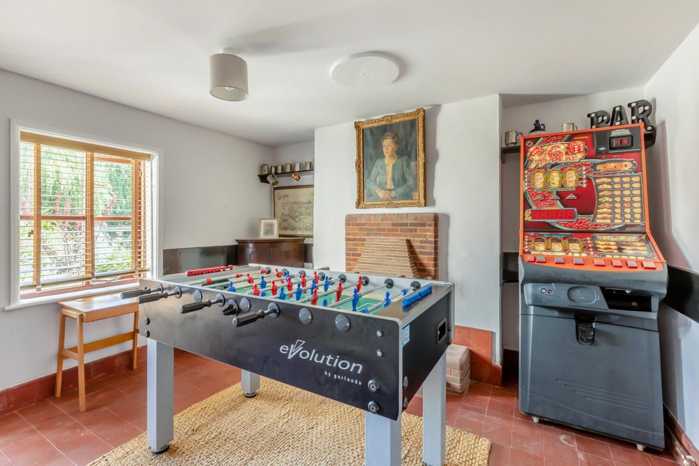 A room with a foosball table, a vintage arcade machine, and a window with blinds.