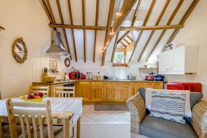 Une cuisine chaleureuse avec des poutres en bois, un coin repas avec une table dressée pour deux personnes, et un espace salon confortable avec un canapé et des coussins.