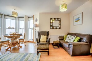 A living room with a brown leather sofa, a wooden dining table, and a chair. There is a lamp, a clock, and a framed picture on the wall. - 2 Bed in Swanage (oc-wy514) (Swanage)