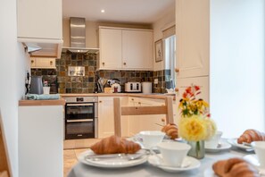 A modern kitchen with a stone backsplash, wooden cabinets, and a dining area with croissants and coffee cups. - 2 Bed in Weobley (oc-c32945) (Weobley)