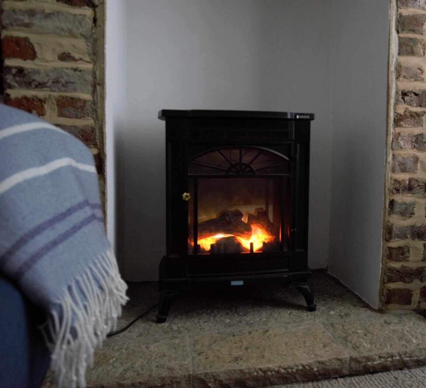 A wood-burning stove in a corner of a room with a brick wall and a blue blanket on a chair.