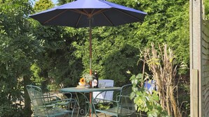 A patio with a table and chairs under a blue umbrella, surrounded by greenery and a wooden fence. - 3 Bed in Leiston (oc-wil) (East Bridge)