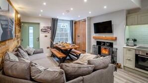 A cozy living room with a sofa, a dining table set with breakfast, a fireplace, and a kitchen area with a TV and modern appliances. - 3 Bed in Whitby (oc-h33106) (Whitby)