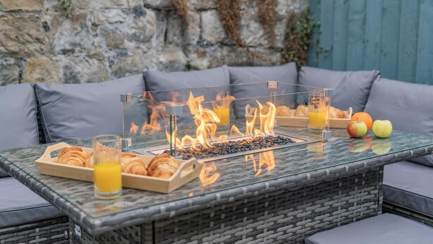 A glass-top outdoor dining table with a built-in fire feature, surrounded by grey cushioned seating. - 3 Bed in Whitby (oc-h33106) (Whitby)