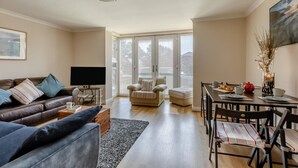 Ein modernes Wohnzimmer mit einer Ledersofa, einem Fernseher, einem Esstisch mit Stühlen und einem großen Fenster mit Jalousien.