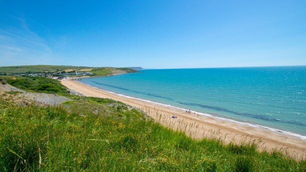 Eine Küstenlandschaft mit Sandstrand, klarem blauen Meer und grünen Hügeln im Hintergrund.