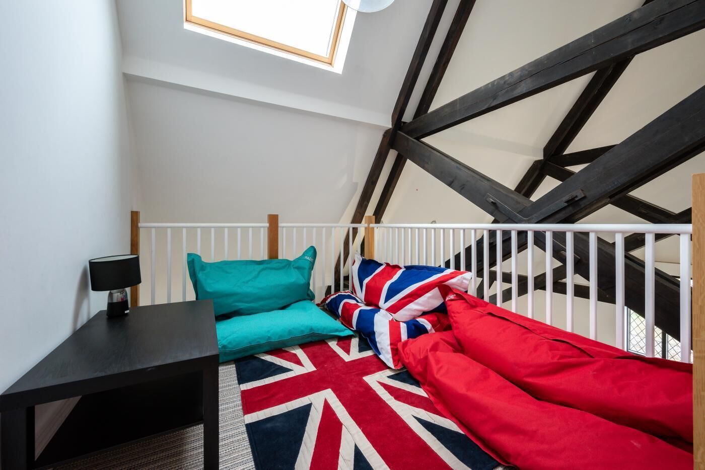 Une chambre à coucher aménagée dans un grenier, avec un plafond haut, des poutres apparentes, une rambarde blanche, un bureau avec une lampe et un lit recouvert d’une couette et de coussins aux couleurs de l’Union Jack.